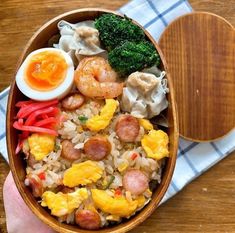 a person holding a wooden bowl filled with rice and veggies next to an egg