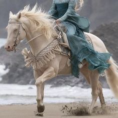 a woman riding on the back of a white horse down a beach next to the ocean