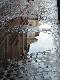 the reflection of a building in puddles of water on cobblestone street with people walking by