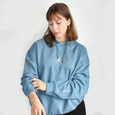 a woman wearing a blue sweatshirt and black pants is looking down at her phone while standing against a white wall