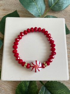 a red and white beaded bracelet with a candy cane charm sits on a table next to leaves