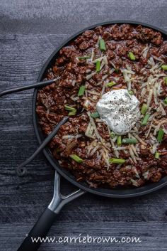 a skillet filled with chili and sour cream on top of a wooden table next to utensils