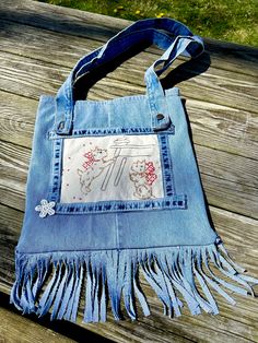 a denim purse with embroidered letters and fringes on the bottom, sitting on a wooden bench