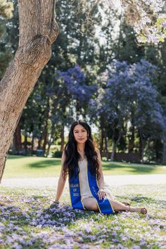 a woman sitting in the grass under a tree with purple flowers around her and looking at the camera