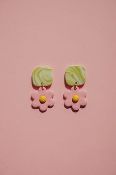 two pink and green flower shaped earrings on a pink surface with yellow accents, one in the shape of a sunflower