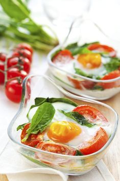 two glass bowls filled with food on top of a wooden table next to tomatoes and green leaves