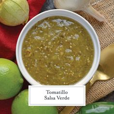 a white bowl filled with green salsa next to limes and garlic on a red cloth