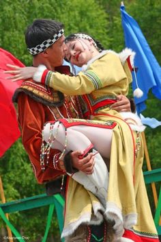 two people dressed in costumes hugging each other on a stage with flags and trees in the background