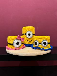 three yellow crocheted hats with eyes and bows on a table next to a pink wall