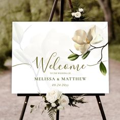 a welcome sign with white flowers and greenery hanging from it's easel