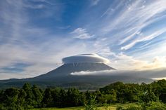 a large cloud is hovering over the top of a mountain with trees in the foreground