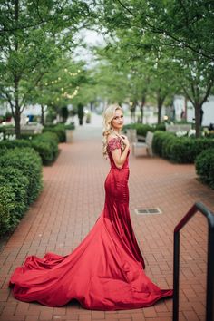 a woman in a long red dress standing on a brick walkway with trees and bushes behind her