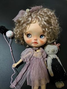 a doll with curly hair and blue eyes holding a stuffed animal in her hand, on a black surface