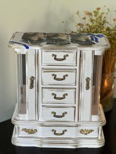 a white and gold painted chest of drawers on top of a black table next to a potted plant