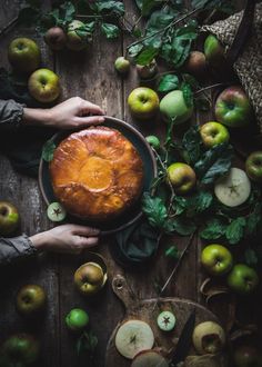 a person holding a pie on top of a wooden table surrounded by apples and leaves