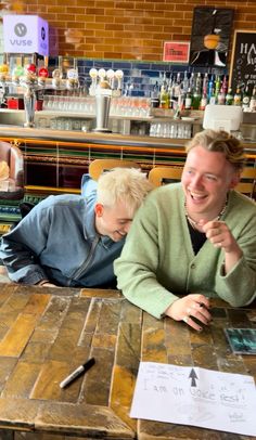 two people sitting at a wooden table with papers on it and one person pointing to the side