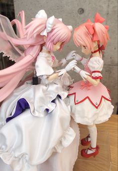 two figurines that are sitting on top of a wooden table, one is wearing a white dress and the other has pink hair