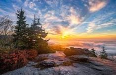 the sun is setting on top of a mountain with clouds and trees in the foreground