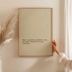 a person holding up a framed quote on a wall next to a vase with some plants