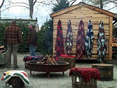 two men standing next to an outdoor fire pit with blankets hanging on the wall behind it