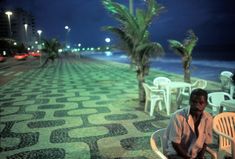 a man sitting on top of a white chair next to the ocean at night with palm trees