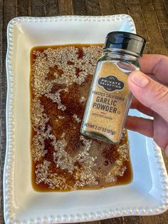 a hand holding a jar of garlic powder on top of a square white plate with wood flooring