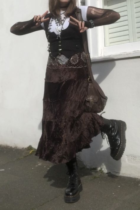 A mid-size person/girl wearing an oversized white fairy crop top, a black waistcoat over it, a black ripped tights top under it, a long dark brown skirt, ripped rose-patterned tights under it, black platform dr. Martens and multiple crystal rings and bracelets. 	The person is also wearing multiple silver necklaces; one in form of a fairy and one in form of a moon, a brown rosary-style and a green bead necklace and a black lace choker. They also have a brown handbag with them. Brown Goth Aesthetic, Maxi Skirt Outfit Grunge, Brown Goth Outfit, Crow Core Aesthetic Outfits, Cottage Goth Fashion, Crow Core Outfit, Steampunk Aesthetic Outfit, Dark Cottage Core Outfits, Fit Aesthetic Outfit