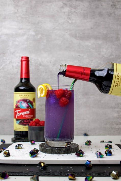 A tall glass is filled with a purplish-blue beverage garnished with raspberries and a lemon twist. There are iridescent crystals scattered on the tray around the glass. A bottle of Torani Galaxy syrup is in the background and another is pouring into the glass. Galaxy Lemonade, Fancy Beverages, Torani Recipes, Syrup Recipes, Butterfly Pea Tea, Twisted Recipes, Lemonade Recipe, Tea Cocktails, Cocktail Mixers