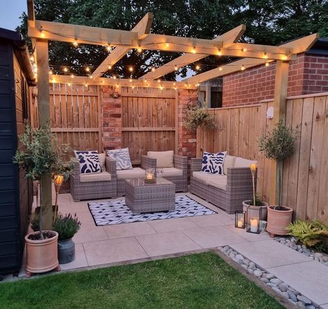 A gray wicker patio set is arranged on a white and black rug laid over beige stone pavers. The patio set is covered by a light wood pergola lit by black festoon lights. Make pergolas cozier with greenery, garden lights, and black lantern dÃ©cor. Garden Patio With Pergola, Fenced In Pergola, Backyard Vision Board, Backyard Deck Pergola Ideas, Garden Ideas Pergola, Pergola In Yard, Garden Design With Pergola, Patio With Fairy Lights, Back Garden Design Ideas