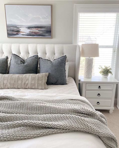 This bedroom features a white tufted bed pushed against the off-white wall underneath a landscape picture. A white bedspread covers the bed on which a light grey coverlet has been tossed. Dark grey pillows and a beige bolster have been placed at the head of the bed. A white bedstand sits on the beige carpet in front of a bright window. Bedroom Ideas Cream Carpet, Cream And Grey Bed, Grey Bed Pillows, Cream And White Layered Bedding, Bed Blanket Arrangement, School House White Bedroom, Grey Bedhead Styling, Gray And Beige Bedding, Grey And White Bedding Ideas