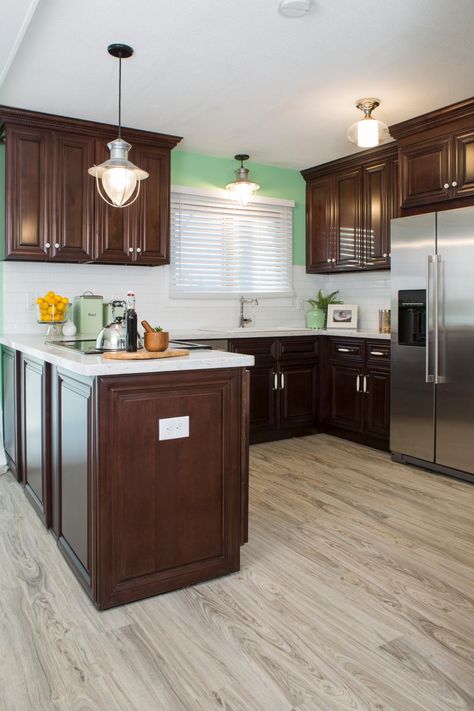 This modern kitchen features dark wood cabinetry paired with mint green walls and a white subway tile backpslash. An industrial pendant light hangs over the stove area, while two additional ceiling fixtures also keep the space well-lit and cheery. Green Wall Brown Cabinet Kitchen, Cherry Cabinets With Light Floors, Dark Cabinet Light Floor, Light Floors With Dark Cabinets, Light Hardwood Floors With Dark Cabinets, Kitchen Flooring With Dark Wood Cabinets, Kitchen Floors With Dark Cabinets, Dark Cabinets With Light Floors, Light Floors Dark Cabinets