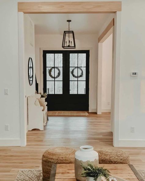 Black front doors create contrast with the white wall and light wood floors of this home's entryway. A black entryway chandelier hangs from the ceiling above a rustic white bench decorated with pillows. Light wood door headers match the wood décor of the rest of the home. Black Entryway Chandelier, Light Wood Door, White Bench Entryway, Stained Front Door, Black Entryway, Wooden Double Doors, Warm Wood Flooring, Front Door Interior, Entryway Chandelier