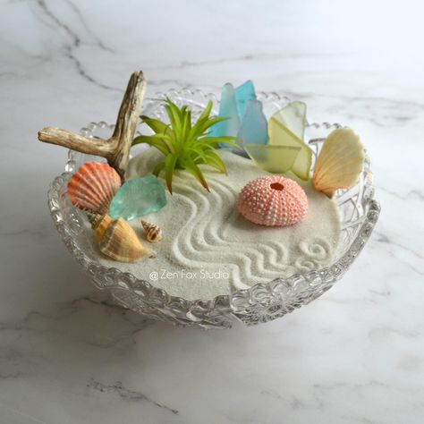 Add some coastal vibes to your space with this Beachy Zen Garden display in a Vintage Imperial Glass Bowl! This one of a kind sand garden sits inside an ornate vintage glass bowl. The air plant is artificial, no maintenance needed. Try adding your own treasures to personalize it!  If you need to relax, rake or trace designs in the sand and rearrange the accessories. Great for display on any desk, bookshelf, or table top.  Includes everything pictured: Imperial Glass Bowl, White Sand, Rake, Bamboo Stylus, 8 Pieces of Sea Glass, 4 Sea Shells, Sea Urchin, Driftwood, Faux Air Plant Base Diameter: 7.75" ★See all of the Zen Gardens available in my shop: https://www.etsy.com/shop/ZenFoxStudio Zen Garden Desk, Sand Boxes, Glass Bowl Centerpieces, Zen Sand, Sand Garden, Shell Mermaid, Succulent Bowls, Diy Succulent Terrarium, Garden Centerpiece