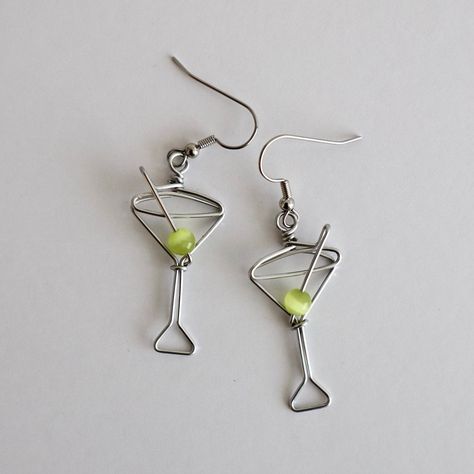 Martini Glass Earrings. Great gift for Bartenders, Hostesses and Waitresses. NOW AVAILABLE IN TWO SIZES. Regular martini glass is 1 1/2 inches high, small martini glass is 1 1/8 inches high (not including earring hooks, see inset photo for scale). Tiny wire martini glasses, complete with green cat's eye bead.  Surgical steel hooks are hypo-allergenic. Clear plastic stoppers on backs. Aluminum wire will not tarnish. I have been making custom wire art and jewelry for 30 years. I started in 1989 selling jewelry on the streets of Montreal. Over the years I have participated in many craft shows, exhibitions and sold my work in gallery shops across Canada including the National Gallery of Canada.  My husband and I specialize in custom wedding cake toppers featuring his wire bicycles and people t Martini Wire Earrings, Wire Wine Charms, Stud Wire Earrings, Wire Earring Designs, Wire Earring Ideas, Unique Diy Earrings Ideas, Wire Earrings Diy, Selfmade Earrings, Wire Jewelry Ideas