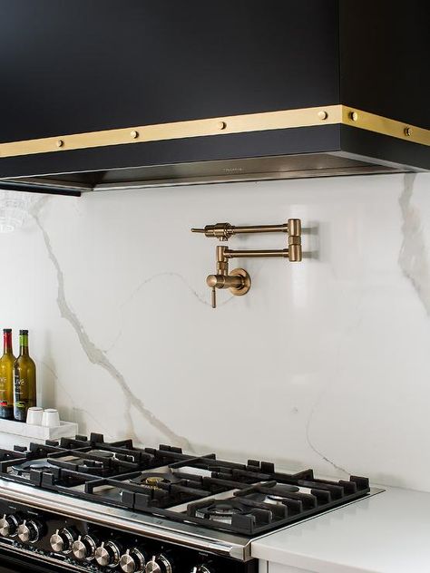 A brass swing arm pot filler is mounted to a marble-look slab backsplash beneath a black and gold dome hood and over a black stove. Black Marble Countertops, Black Stove, White Wood Paneling, Kitchen Island Tops, Pot Filler Kitchen, Quartz Backsplash, Stainless Steel Stove, Kitchen Hood, White Kitchen Island