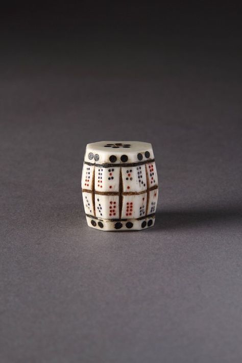 An ivory playing dice shaped to look like a barrel. It has eight sides angled upwards and eight sides angled downwards, separated horizontally by 3 thin brown bands - two near the top and bottom and one cutting across the middle. Each side is marked with a constellation of spots in a combination of red and black colours. Ancient Games, Ok Bye, Dice Collection, Gaming Dice, Ancient Queen, Shape Games, Vintage Board Games, Game Dice, Tabletop Games