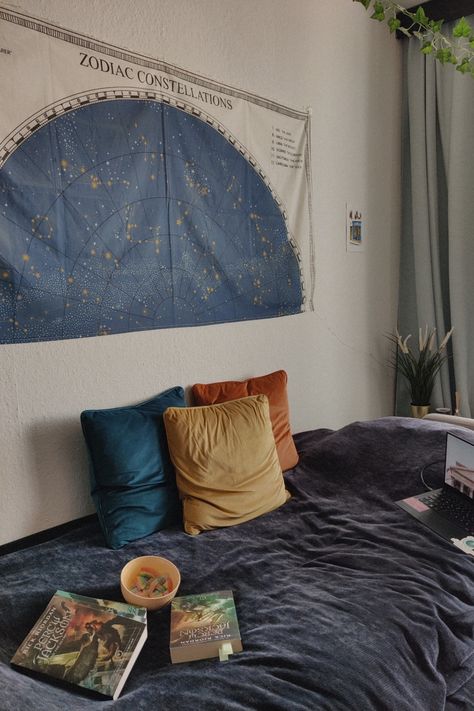 A bed covered with a dark blue velvet blanket, three yellow, blue and orange pillows, the Percy Jackson books, a computer and a yellow bowl with rainbow candy. On the white wall is a tapestry of the zodiac signs and a painting of a parisian street. From the ceiling hangs a vine and the curtains are light blue. Behind the bed is a plant in a golden pot and a red candle light. Astrology Dorm Room, Percy Jackson Themed Bedroom, Aquarius Bedroom Aesthetic, Percy Jackson Room Aesthetic, Astrology Themed Bedroom, Percy Jackson Bedroom Ideas, Percy Jackson Room Ideas, Room Inspo Vines, Percy Jackson Decor