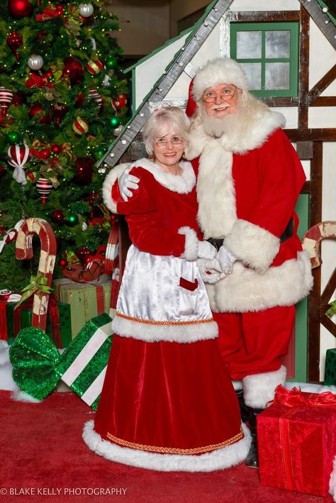 Santa and Mrs. Claus standing in front of a small green elf house next to a Christmas tree. Both are wearing red and smiling. Mrs Clause Photoshoot, Female Santa Claus, Santa Claus Wife, Santa And Mrs Claus Photo Session, Father Christmas Costume Santa Suits, Mrs Claus Outfit, Mrs Santa Claus, Santa Claus Pictures, Mrs Clause