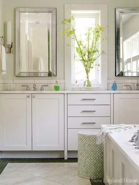 White bathroom cabinets and white countertops.  A slim white tiled backsplash accents the vanity whilst modern polished nickel faucets add sparkle. Above the drawers and in front of the window, an inlaid bone tray holds a vase with a bright spray of yellow flowers. Next to the paneled bath surround an inlaid stool with a minty green colorway adds pattern. The border around the floor is a deep green mosaic tile with an inset marble tiled center layered at an angle to expand the space Bathroom Vanity Window In Middle, Double Vanity Window In Middle, Windows Above Bathroom Vanity, Master Bath With Window Between Sinks, Window Between Vanity Sinks, Bathroom Window Above Sink, Bathroom Vanity With Window In Middle, Window Above Vanity In Bathroom, Vanity With Window In Middle