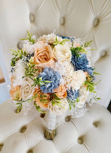 This bouquet features pretty sola flowers in shades of dusty blue, peach, and ivory. Faux greenery and preserved babies breath add soft accents. Pictured bouquet is 10 inches in diameter. Coral Blue And White Wedding Flowers, Dusty Blue And Peach Wedding Cake, Spring Wedding Color Palette 2023, Blue And Peach Bouquet, Dusty Blue And Peach Wedding Theme, Dusty Blue And Peach Wedding, Peach And Blue Wedding, Blue And Peach Wedding, Blue Peach Wedding