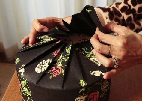 Instead of finding a rectangular box for a round package like a cookie tin, try the fashion trick of pleating. Warning to the easily flustered: This takes practice. For a round cookie tin, Sumika used  Cream & Red Roses on Black  gift wrap, a striking and elegant alternative to traditional reds and greens.       VIDEO: Wrapping round presents      She cut a piece as wide as the circumference of the circle (plus a half-inch extra) and as long as the diameter of the lid plus one-half the height o… Japanese Gift Wrapping, Gift Wrapping Techniques, Wrapping Presents, Gift Wrapping Ideas, How To Wrap, Japanese Gifts, Creative Gift Wrapping, Diy Gift Wrapping, Present Wrapping