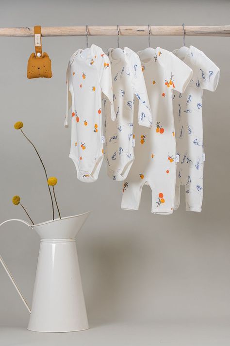 Four baby body suits hanging on a rail in an all-over Clementine print and a Blue Shrimp print made from GOTS certified organic cotton styled with a hanging orange brown sleeping bear rattle next to a tin watering can containing 3 yellow flowers. Baby Product Photography, Baby Clothes Photography Ideas, Kidswear Product Photography, Flat Lay Photography Kidswear, Swaddle Product Photography, Baby Store Display, Kids Clothing Store Design, Playful Blue Cotton Onesie, Playful Organic Cotton Onesie For Playtime