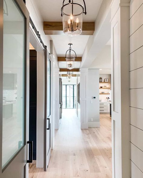 As you enter this front door, you will notice the white walls, a beige barn door, and a warm wood floor. A series of glass pendant lights are attached to the white ceiling. Ponder installing warm wood beams on the ceiling for added dimension. Light Blue Hallway, Accent Ceiling Ideas, Hallway Pendant Lighting, White Metal Bed, Hallway Chandelier, White Hallway, Warm Wood Flooring, Chandelier Ideas, White Wainscoting