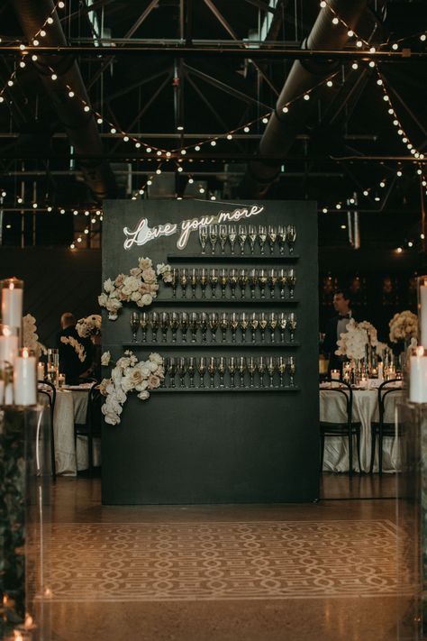 This image features a black champagne rack used for a wedding reception and cocktail hour decor. It has two floral arrangements with blush and white roses and white orchids. It also features a love you more neon sign. Champagne And Black Wedding Theme, Wedding Reception Cocktail, Blush Wedding Reception, Wedding Reception Cocktail Hour, Cocktail Hour Decor, Reception Cocktail, Divine Union, Indian Reception, Drink Display