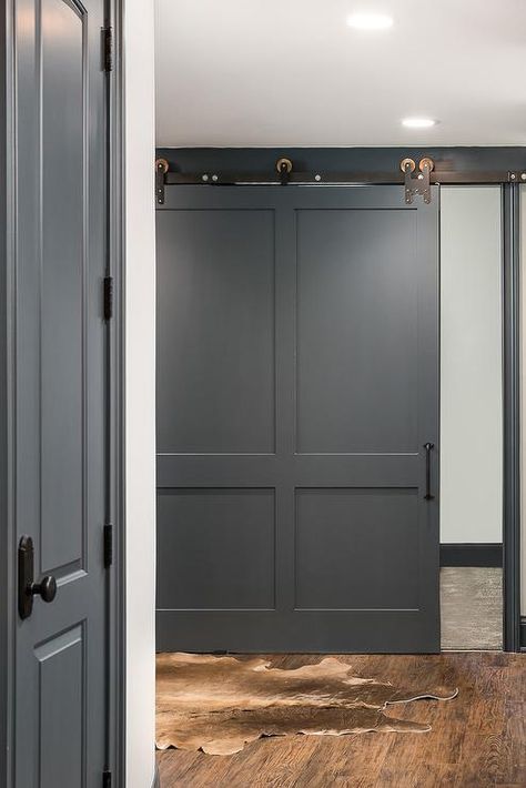 A charcoal gray barn door on rails opens to a basement boasting a brown cowhide rug. Raccoon Fur Paint Color, Dark Grey Baseboards, Raccoon Fur Benjamin Moore, Benjamin Moore Gunmetal, Wolf Gray Benjamin Moore, Benjamin Moore Raccoon Fur, Transitional Basement, Grey Painted Furniture, Room Ideas Dark