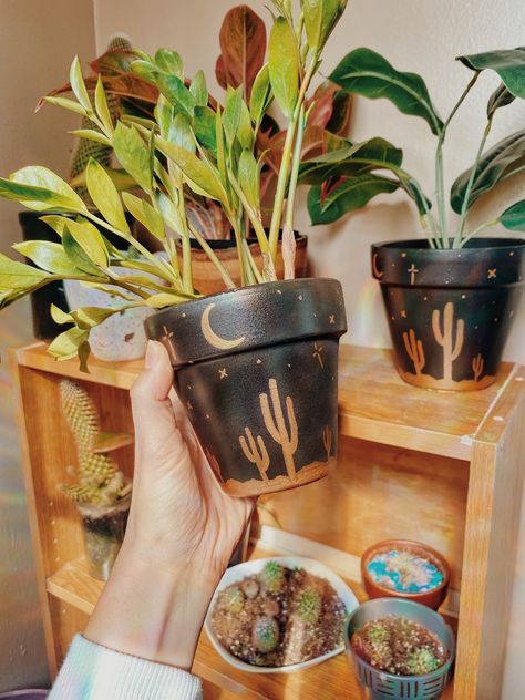 "✨Our Desert Sky Pot is sure to make a statement for all your plant decor dreams!✨ ✨This design is handpainted on standard terracotta clay style pottery. Beautiful sleek black with a metallic gold saguaro desert scene to finish the look.  SIZES: 4\" pot, 6\" pot, 8\" pot ✨All pots have drainage holes & sealed with clear enamel gloss for a chip-free aesthetic! NOTE: each design is hand-painted, so please allow room for the creative process as no two items are exactly alike" Boho Painted Plant Pots, Diy Plant Pot Ideas, Diy Terracotta Pots Paint Ideas, Painted Clay Pots Ideas, Planter Painting Ideas, Painted Pots Diy Terra Cotta, Diy Painted Flower Pots, Terracotta Pots Painted, Hand Painted Plant Pots