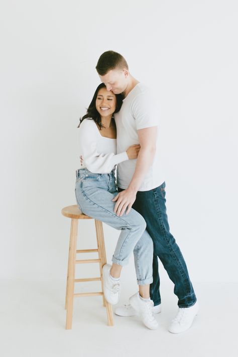 Couple poses while girl sits on a stool and guy hugs her from the side. Stool Couple Photoshoot, Photoshoot With Stool Outside, Couple Shoot Poses Studio, Couple Studio Photoshoot Ideas Simple, Couples Jeans Photoshoot, Jean Couple Photoshoot, Blue Jean White Shirt Couple Photoshoot, Couples Photoshoot White Background, White Shirt Couple Photoshoot