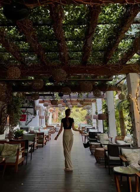 Girl photographed from the back, full body is visible and takes up one third of the vertical space, girl is wearing white pants and a black cropped T-shirt, a handbag is swinging on the right arm creating the illusion of the girl turning around. Girl has long hair tied in a low pony. The ceiling of the restaurant is a wood installation to hold up the vines. On the left and right are seats with green pillows, adding to the overall green tones of the picture Marbella Aesthetic Night, Marbella Nightlife, Spain Aesthetics Malaga, Spain Streets Aesthetic, Greens Restaurant, Marbella Old Town, Marbella Spain, Marbella, Spain