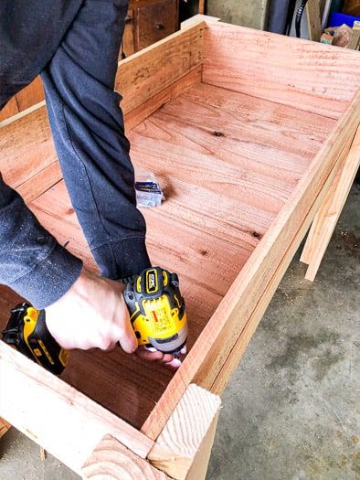 Inside of a raised garden bed showing a person holding a drill to screw in the slats on the bottom Waist High Raised Garden Beds Diy, Bed On Stilts, High Raised Garden Beds, Build A Raised Garden Bed, Wooden Garden Bed, Raised Planter Beds, Building A Raised Garden, Raised Garden Beds Diy, Raised Planter