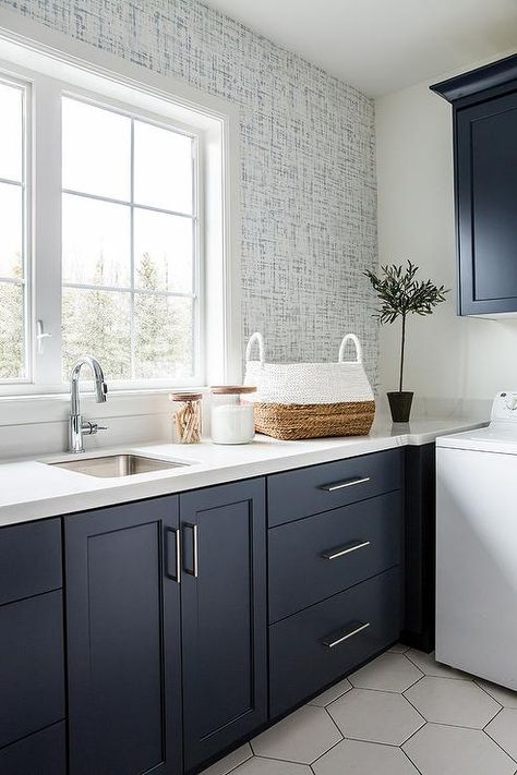 White and blue wallpaper binds together colors and tones in a transitional laundry room featuring dark blue cabinets and large white hexagon floor tiles. Blue Cabinets Laundry Room Ideas, Floor And Decor Hexagon Tile, Laundry Room Ideas Dark Cabinets, Navy Blue And White Laundry Room, White Hexagon Kitchen Floor, Dark Blue Utility Room, Laundry Room With Blue Cabinets, Blue Kitchen Cabinets Tile Floor, Blue Cabinets With Wallpaper